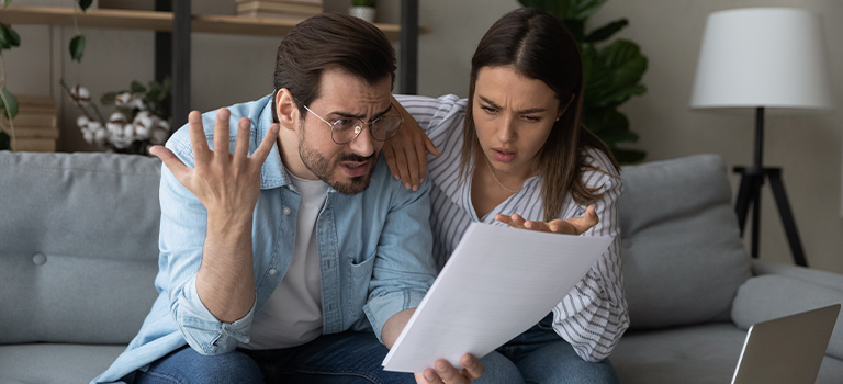 A stressed young couple sitting on a sofa while anxiously reviewing a document together.